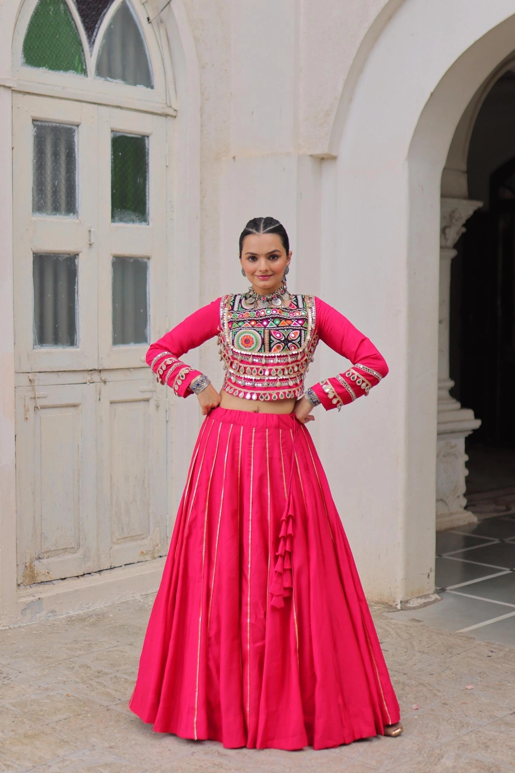Woman in a traditional outfit with a pink top and red skirt standing in front of a white archway.